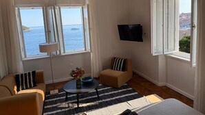 A room with a sofa, armchair, and a small table with a vase of flowers. There is a TV mounted on the wall and a window with a view of the sea. - Onyx Apartments 2&4 (Dubrovnik)