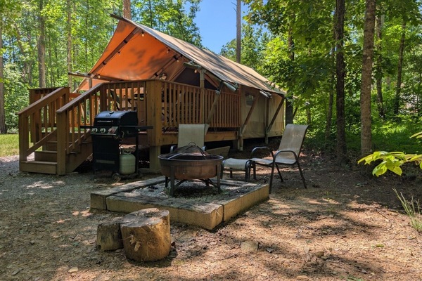 The deck stairs lead up; the fire pit sits just below. Propane grill at left, two chairs around the ring, stumps for extra seating. Most of your evenings happen right here.