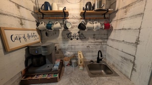 A compact kitchenette with a coffee maker, mugs, a sink, and a shelf with kitchenware.