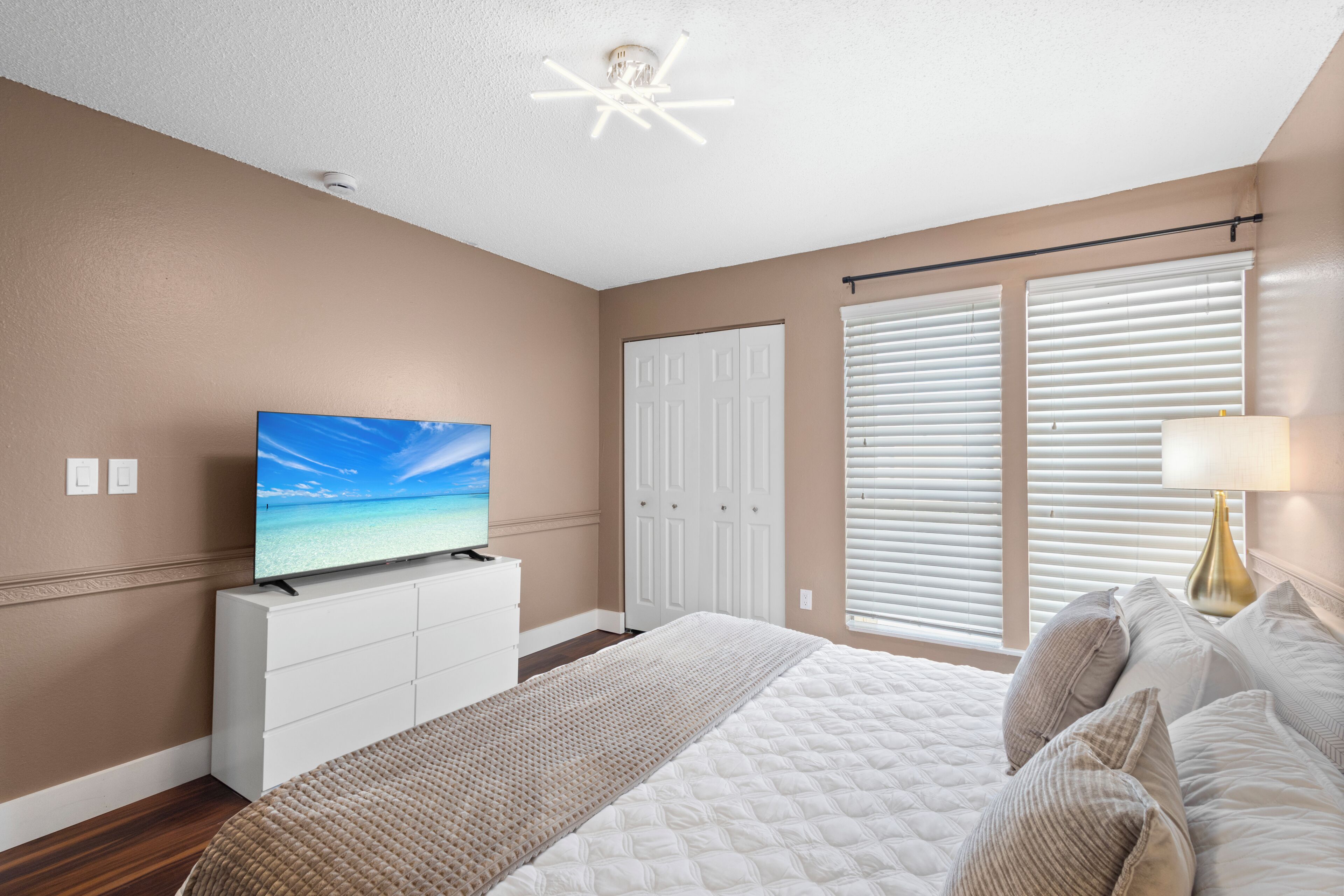Una camera da letto con un letto, una TV su una cassettiera, una finestra con persiane e una luce a soffitto.