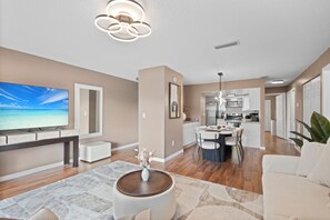 A modern living room with a flat-screen TV, a dining area, and a kitchen in the background. - New 3BR , Heated Pool , 5 Min to Siesta (Sarasota)