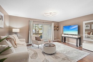 A modern living room with a sofa, armchair, coffee table, TV, and a large window with curtains. - New 3BR , Heated Pool , 5 Min to Siesta (Sarasota)
