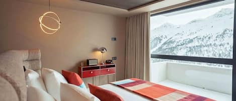 Habitación de hotel con cama, mesita de noche, lámpara, vistas a montañas nevadas y un balcón.