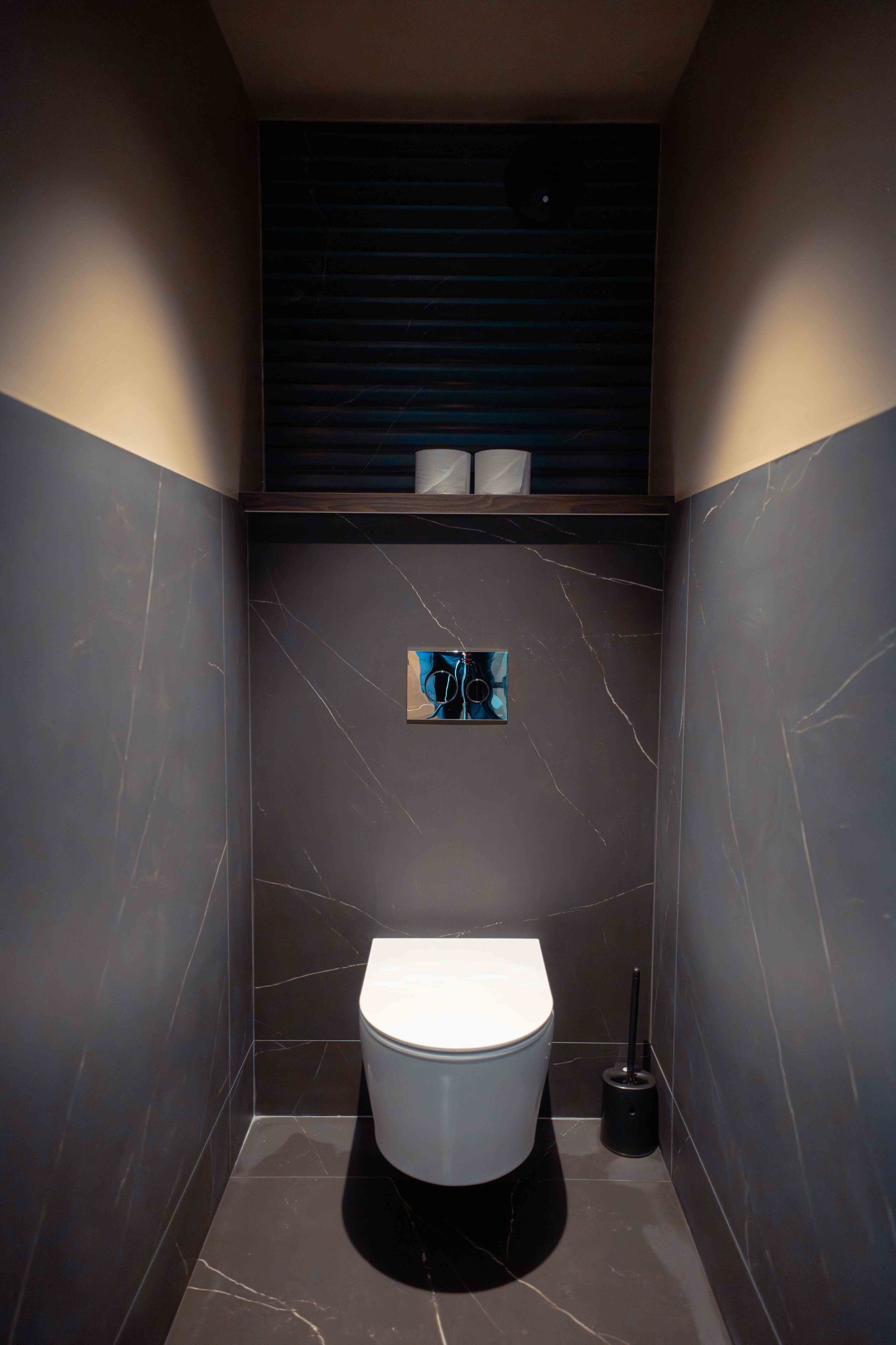 Une salle de bain moderne équipée de toilettes, d’un porte-rouleau de papier toilette fixé au mur et d’une brosse de toilette posée au sol.