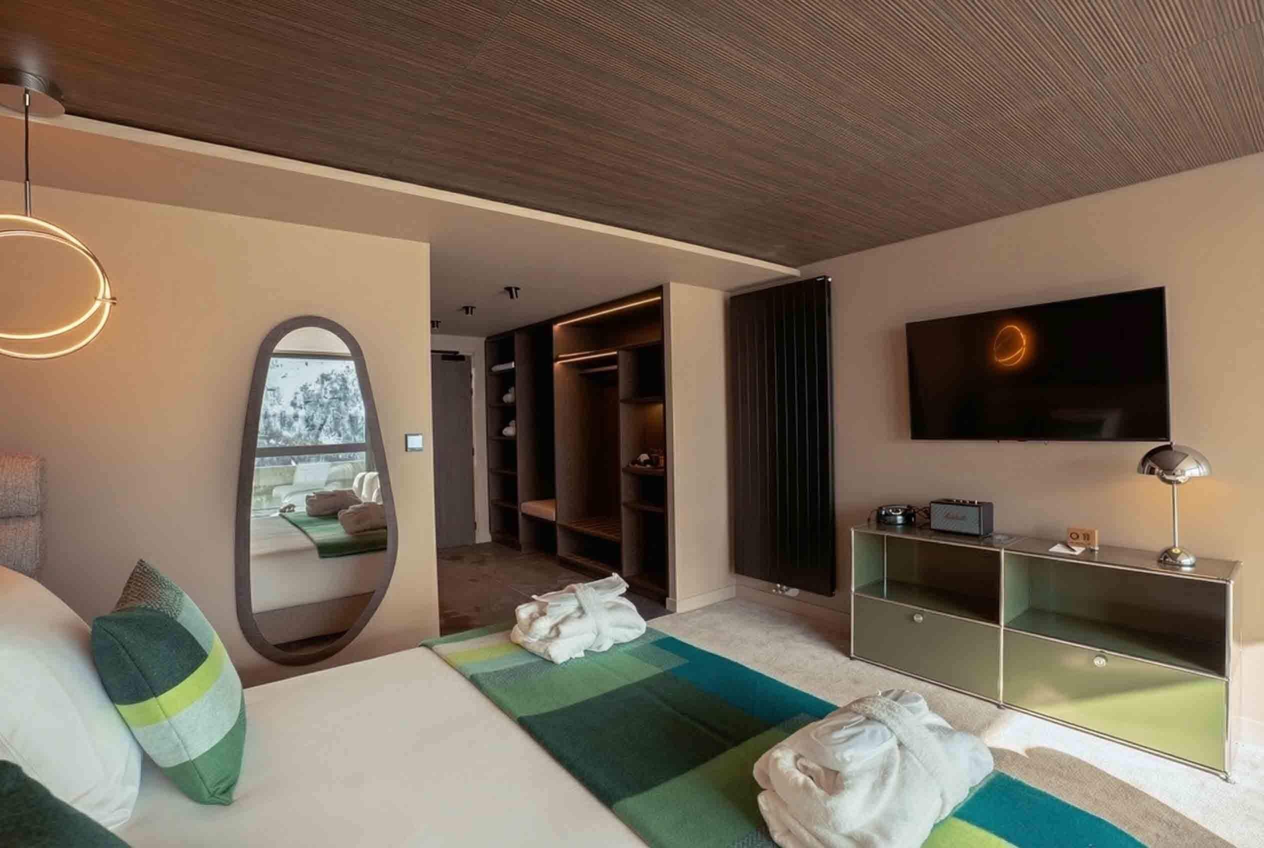A modern hotel room with a large bed, a mirror, a TV, a walk-in closet, and a view of the outdoors.