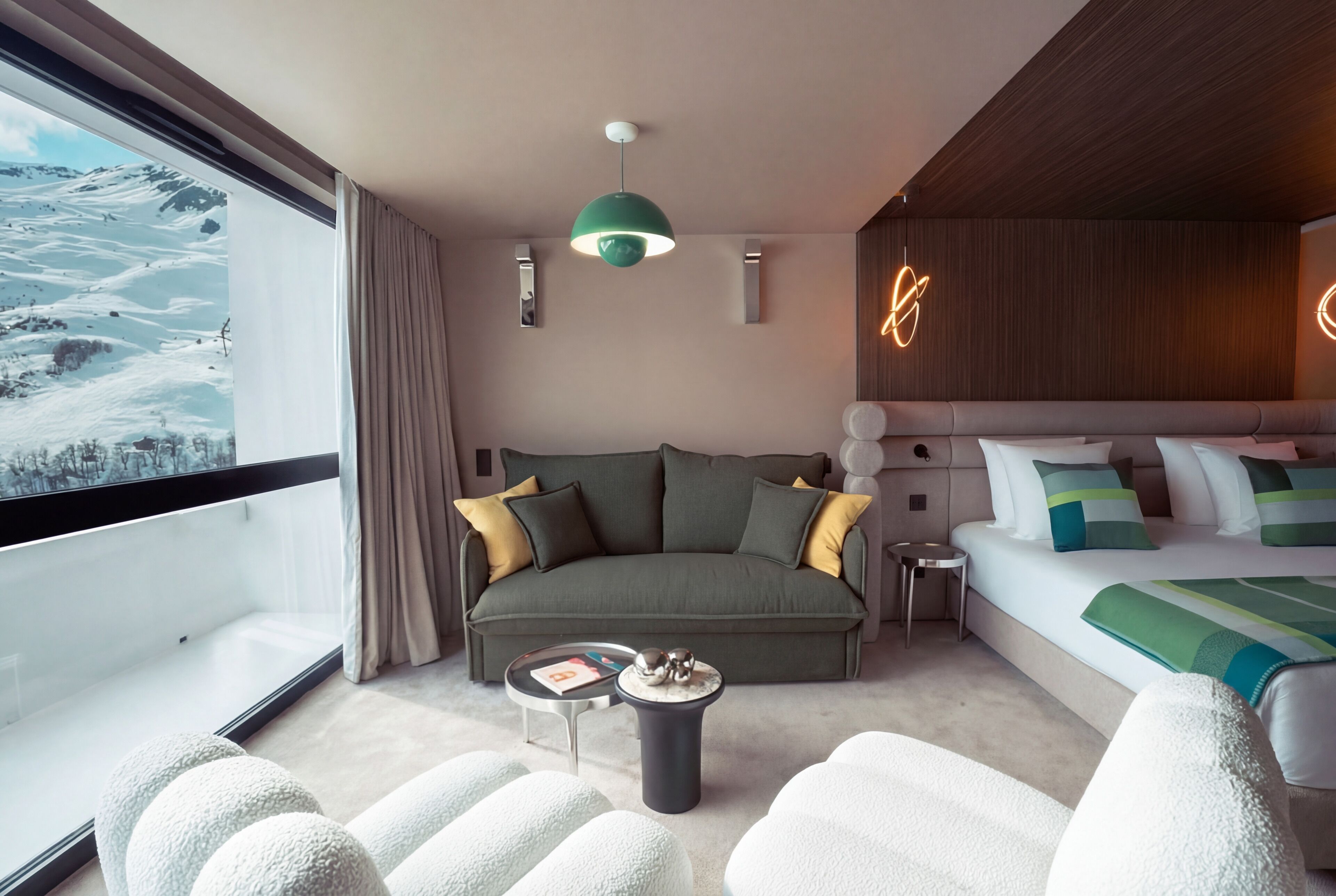 A modern hotel room with a large window, a sofa, a bed, a round table, and a unique ceiling light.