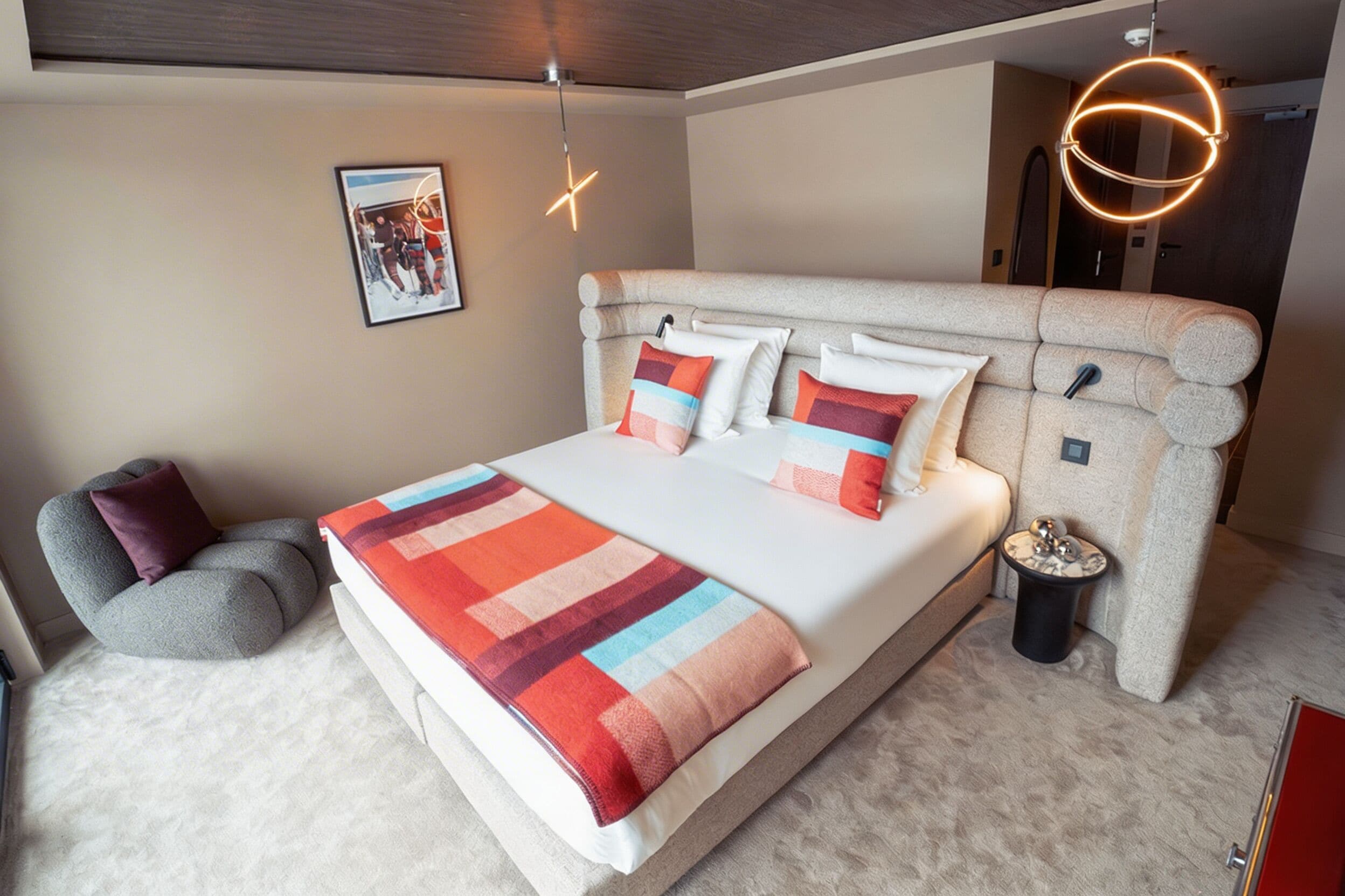 A bedroom with a large bed, a headboard, a bedside table, a chair, a picture on the wall, and a unique circular light fixture.
