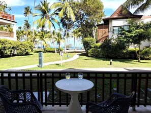 A balcony with two chairs and a table, overlooking a well-maintained lawn with palm trees and a building in the background. - Gorgeous large condo, quiet beachfront complex (Las Terrenas)