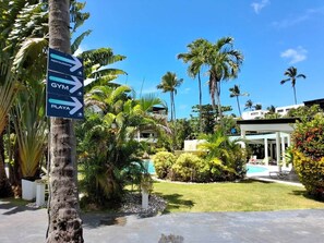 A signpost with directions to a restaurant, gym, and pool area, surrounded by tropical vegetation and a modern building. - Amazing two bedroom Penthouse at Aligio Aparthotel (Las Terrenas)