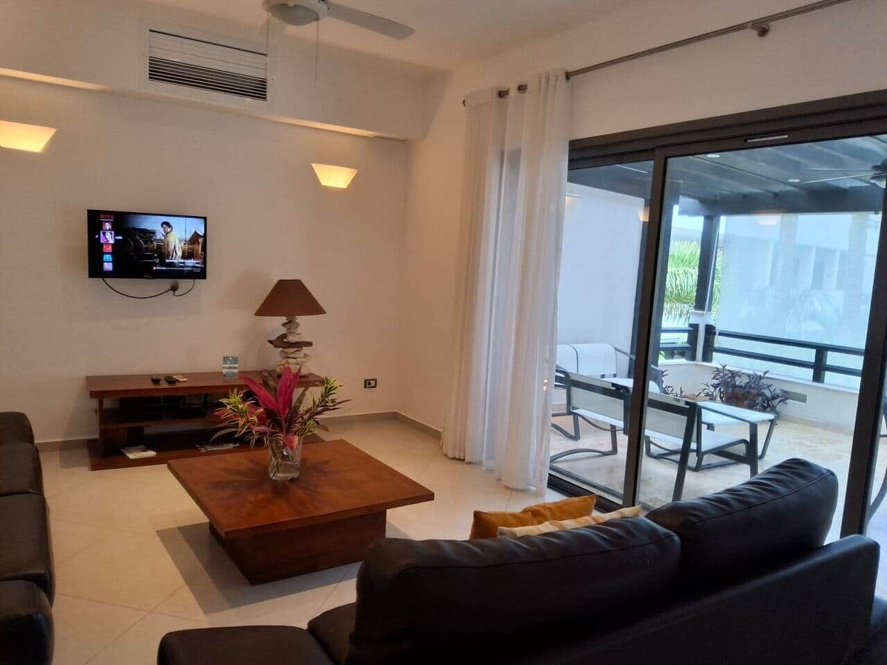 A modern living room with a flat-screen TV, a coffee table with a plant, a sofa, and a balcony with outdoor furniture.