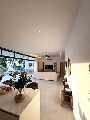 A modern living room with a large TV, white sofas, a dining area with a table and chairs, and a view of the ocean. - Stunning condo, private rooftop pool, best in LT (Las Terrenas)