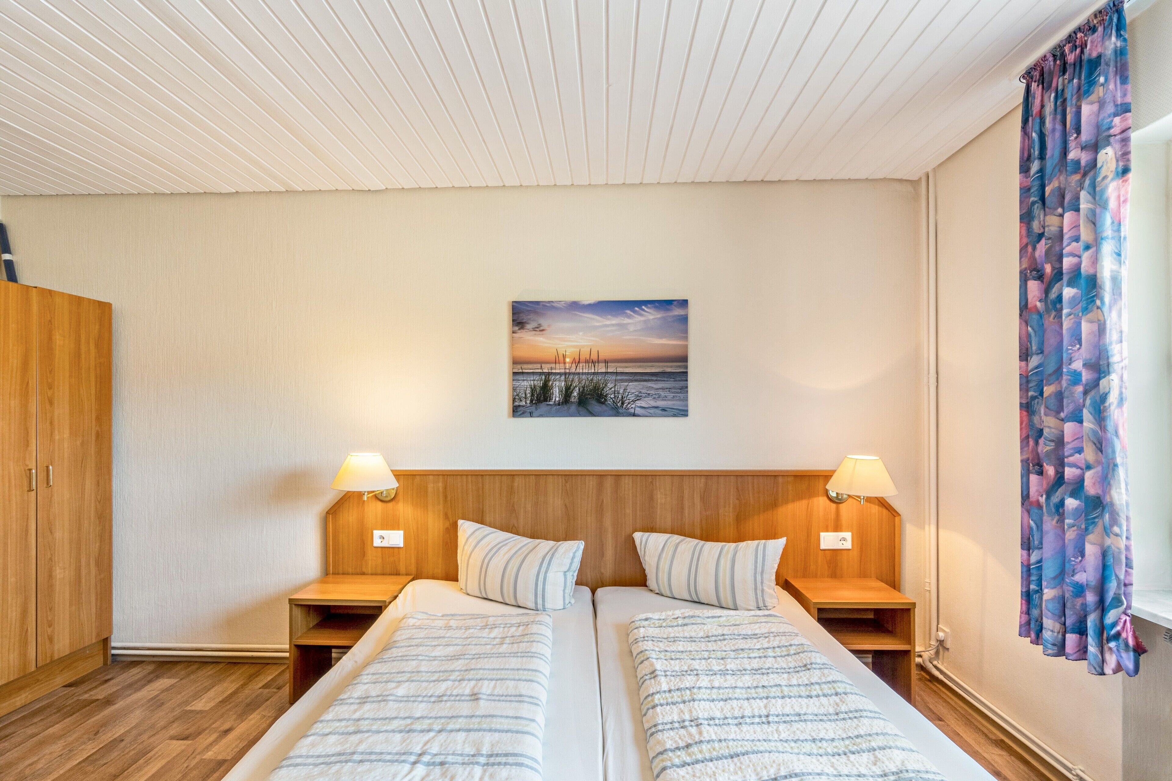 Une chambre double avec deux lits, une tête de lit en bois, des lampes de chevet, un tableau au mur et une fenêtre avec des rideaux.