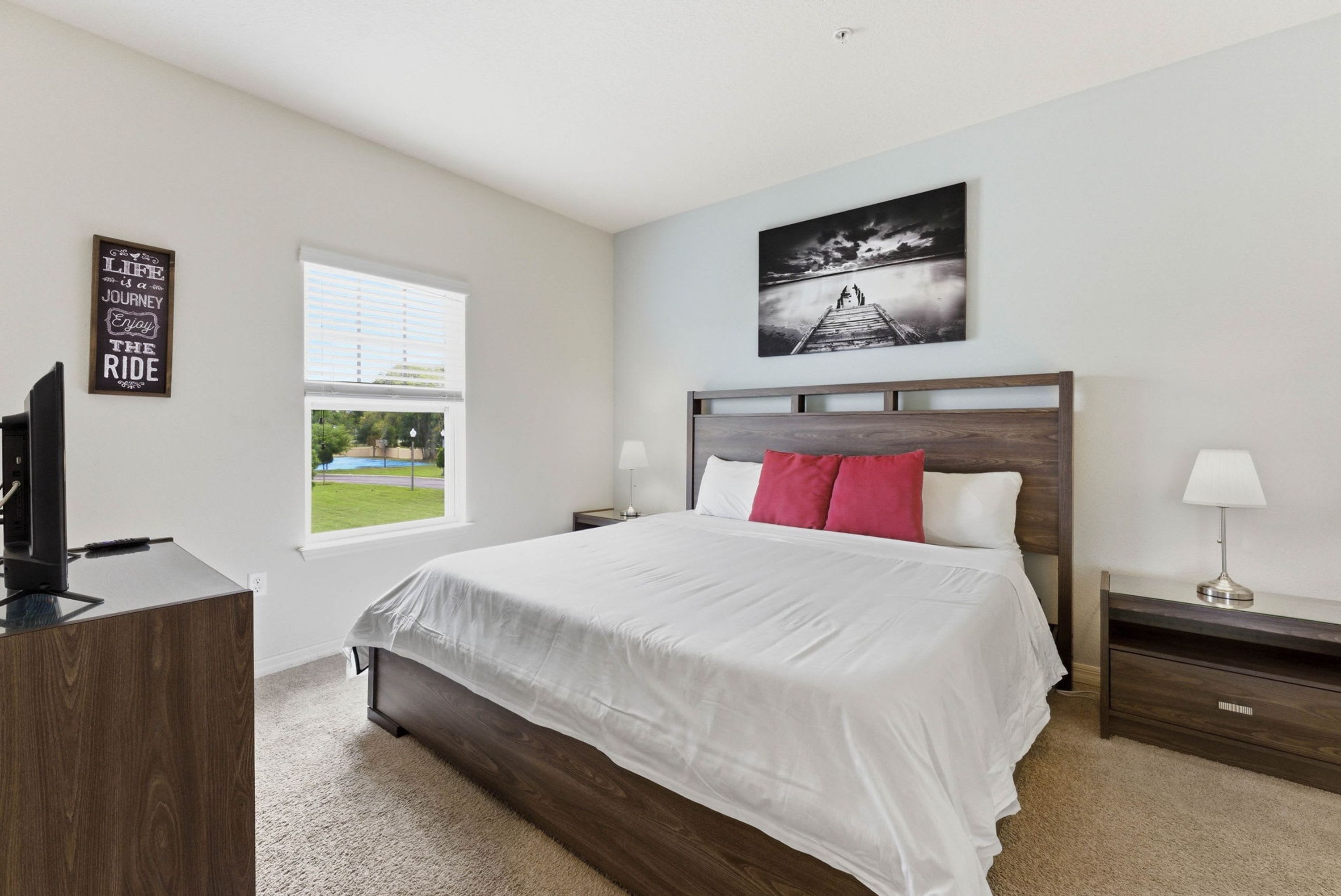 A bedroom with a bed, a desk, a window, a nightstand, and a wall with a picture and a sign.