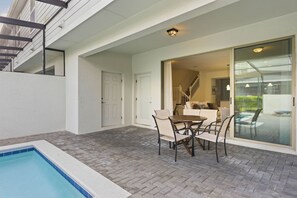 Eine Poolterrasse mit Tisch und Stühlen, eine Glastür zu einem Innenbereich und eine Treppe zu einer oberen Ebene.