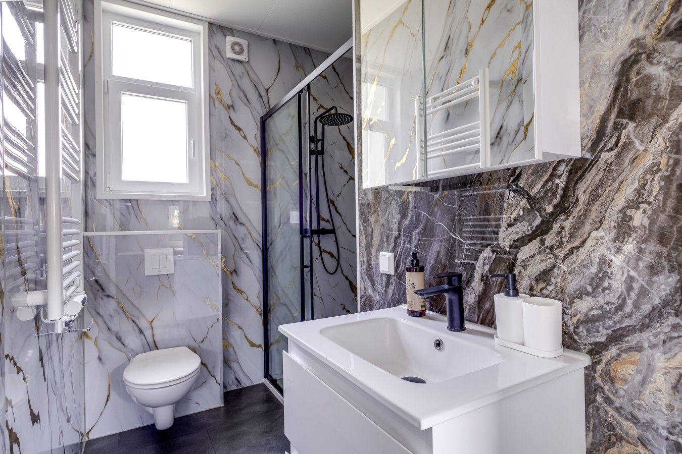 A bathroom with a toilet, shower, and sink, marble walls, and a window.