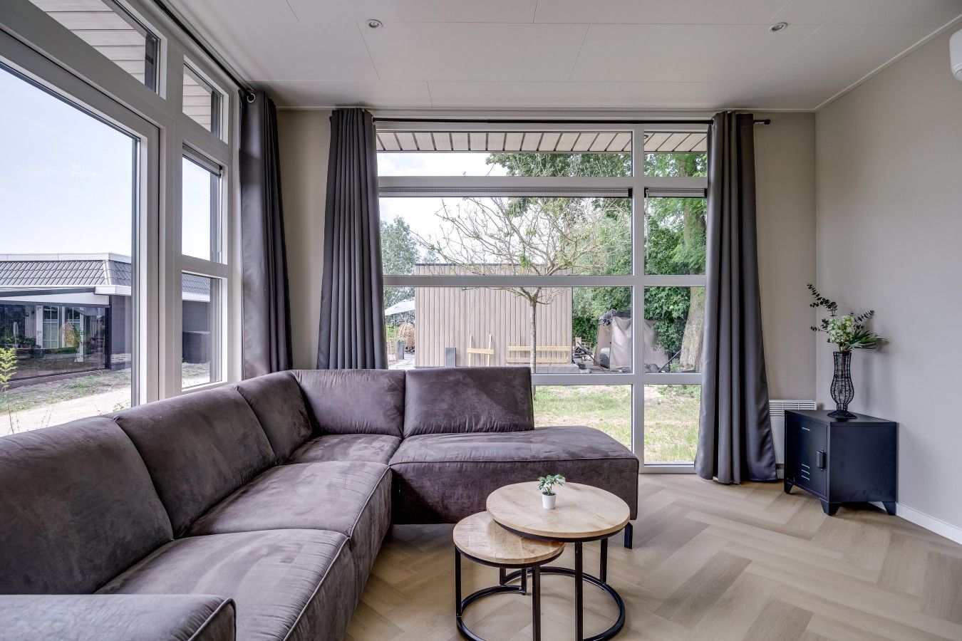 A modern living room with a sectional sofa, a round wooden table, and a view through large windows to the outdoors.