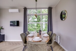A dining area with a table set for four, a television on the wall, and a window with curtains. - Lodge 79 | 6 persoons chalet in Zennewijnen (Zennewijnen)