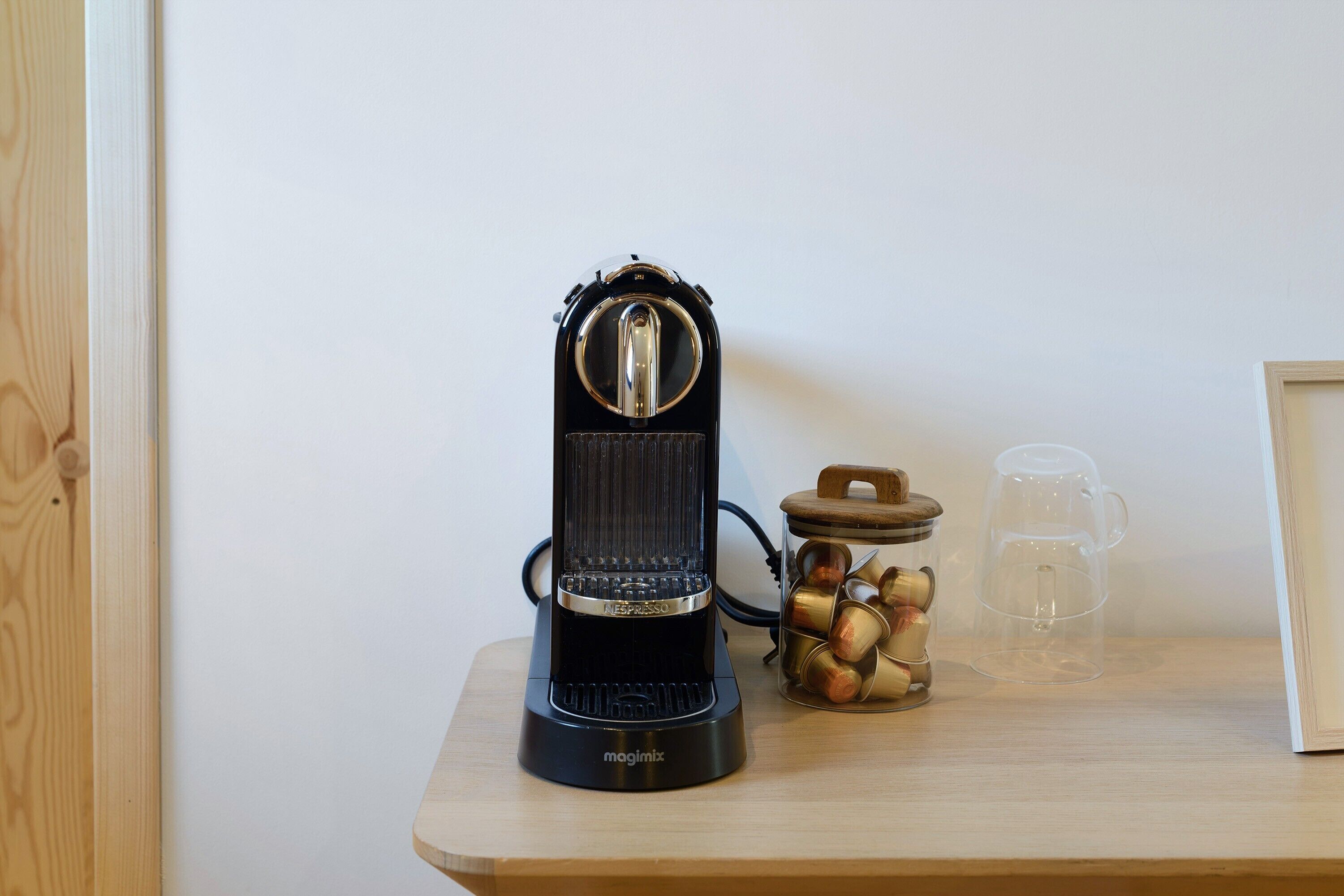 Una macchina da caffè espresso nera su un tavolo di legno, con un barattolo di capsule per caffè e una tazza di vetro.