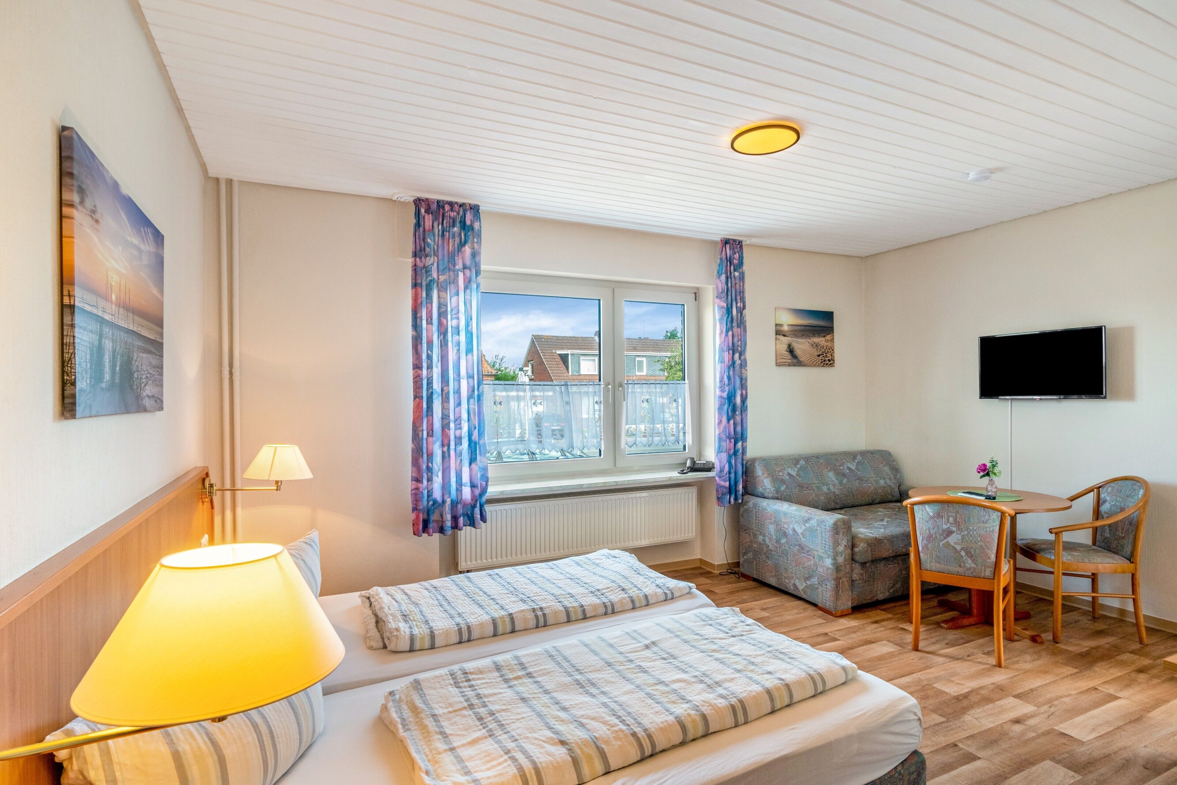 Ein Hotelzimmer mit einem Bett, einer Couch, einem Sessel, einem Tisch, einer Lampe, einem Fernseher und einem Fenster mit Vorhängen.