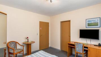 Habitación de hotel con cama, dos sillas, escritorio con computadora, televisor y puerta con cartel de no fumar.