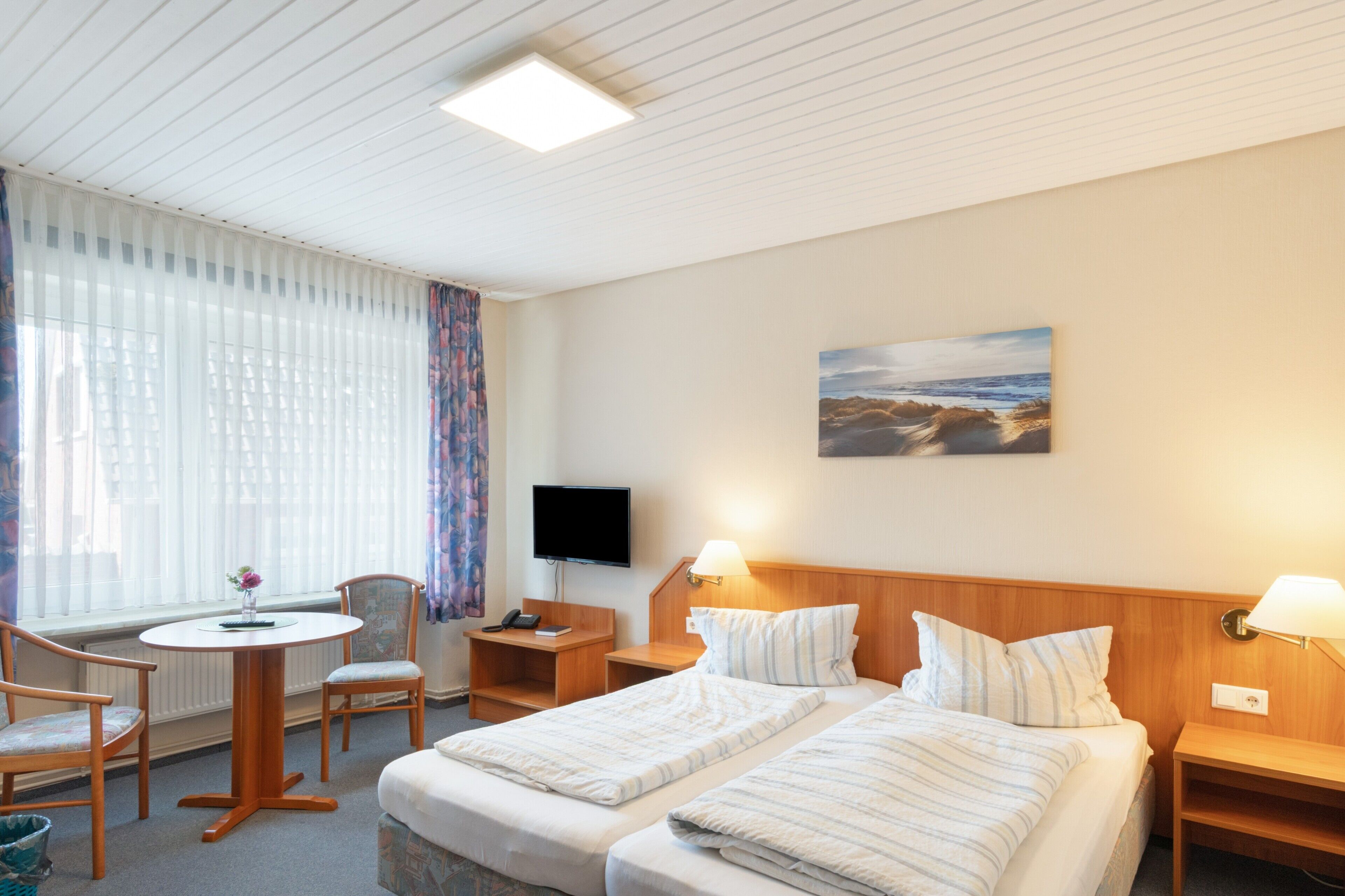 Habitación de hotel con cama, mesitas de noche, televisor, escritorio con silla y ventana con cortinas.