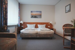 Ein Hotelzimmer mit einem Doppelbett, zwei Nachttischen mit Lampen, einem Sessel und einem kleinen Tisch mit einer Vase. An der Wand hängt ein Gemälde.