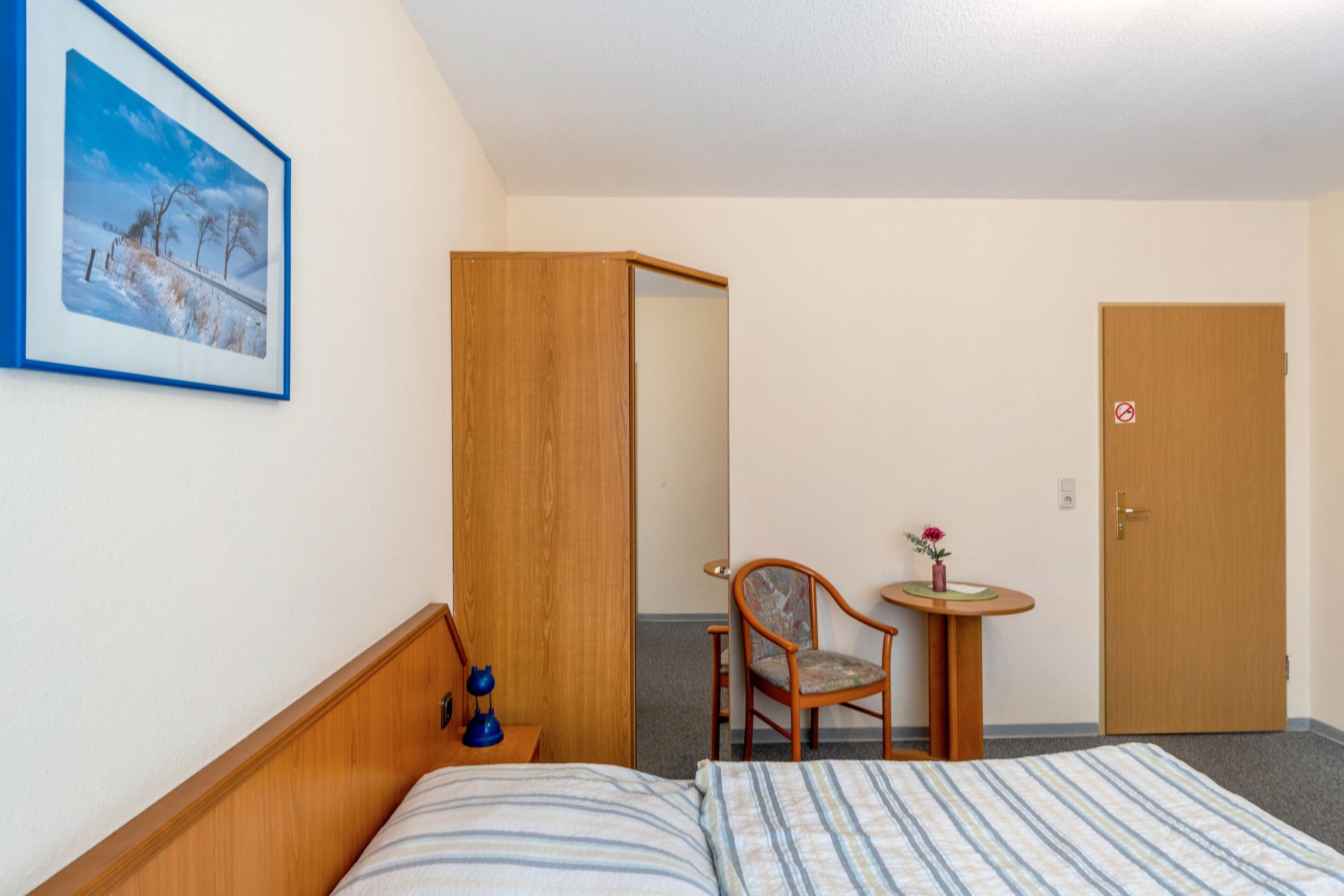 Ett hotellrum med en säng, en stol, ett litet bord med en vas, en garderob i trä och en dörr med en skylt som säger ”rökning förbjuden”.