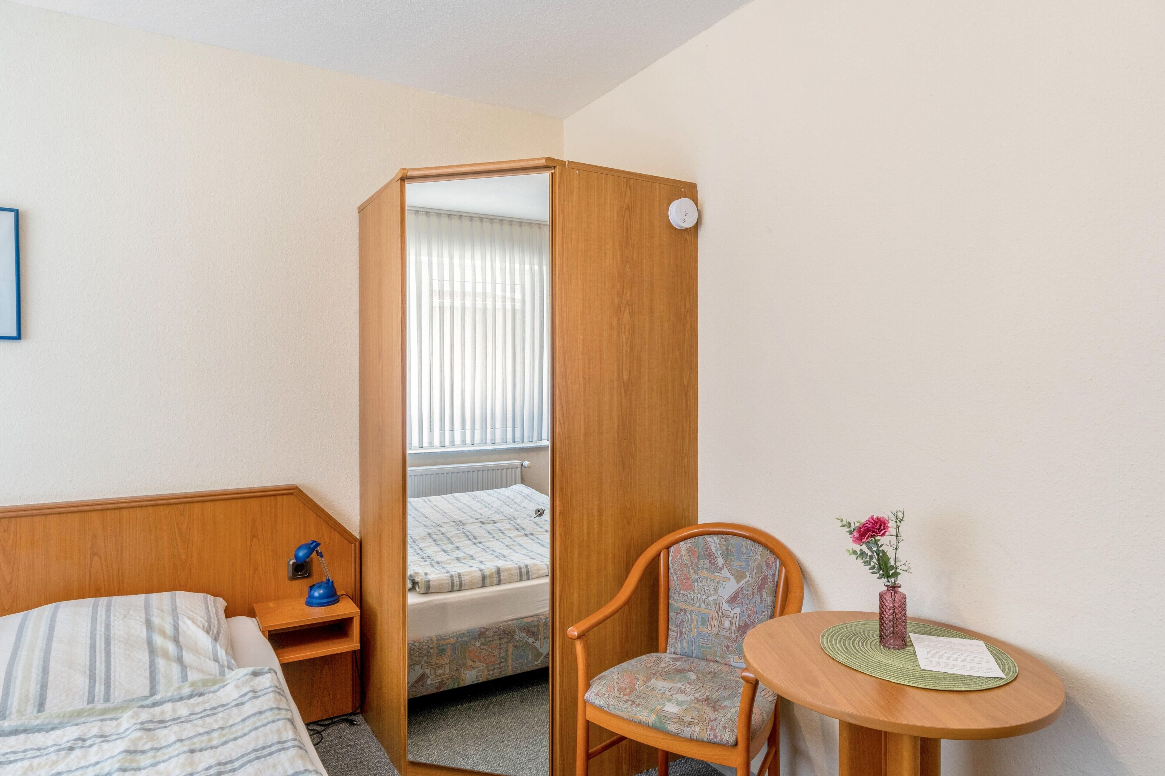 A hotel room with a bed, a desk with a lamp, a chair, a small table with a vase, and a window with blinds.