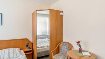 A hotel room with a bed, a desk with a lamp, a chair, a small table with a vase, and a window with blinds.