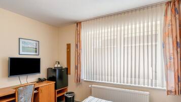 A hotel room with a bed, a desk with a computer, a chair, a TV, and a window with curtains.