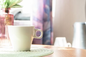 Una taza blanca sobre una mesa de madera con un mantelito verde, un florero rosa con una planta verde y una taza blanca al fondo.
