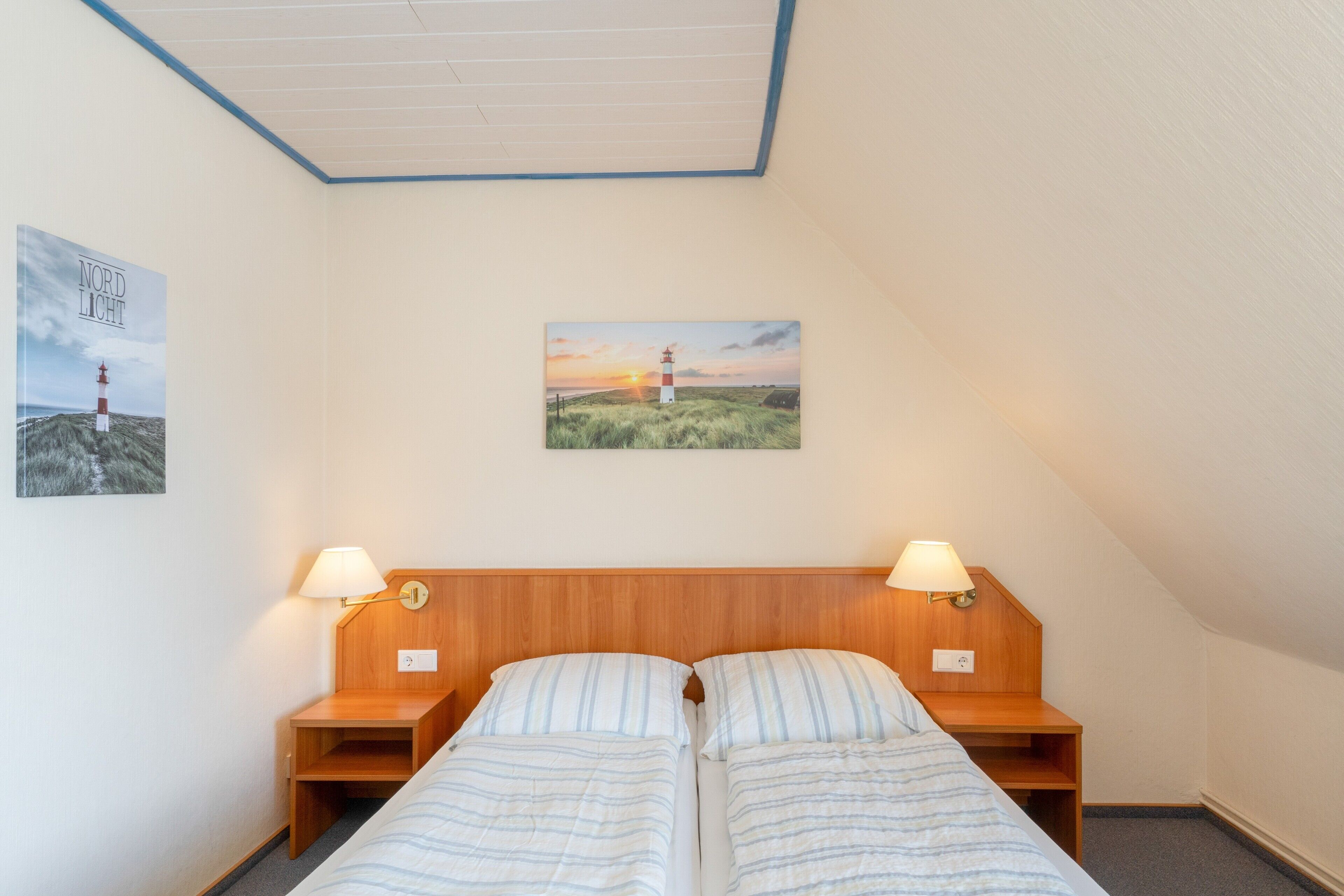 Um quarto com duas camas individuais, cabeceira de madeira, mesas de cabeceira com luminárias, um quadro na parede e teto com detalhes azuis.