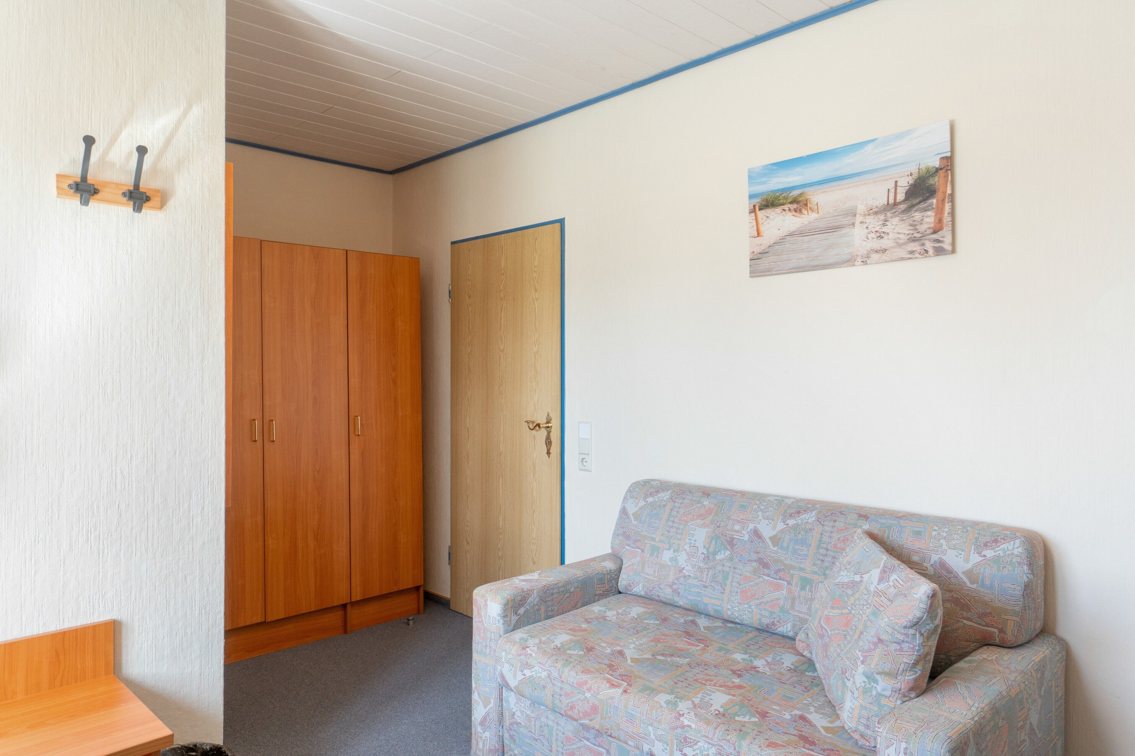 Ein Zimmer mit einem Holzkleiderschrank, einem gemusterten Sofa, einer Tür und einem Wandbild.
