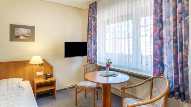 Une chambre d’hôtel comprenant un lit, un bureau avec une lampe, une télévision, une fenêtre avec des rideaux et une chaise.