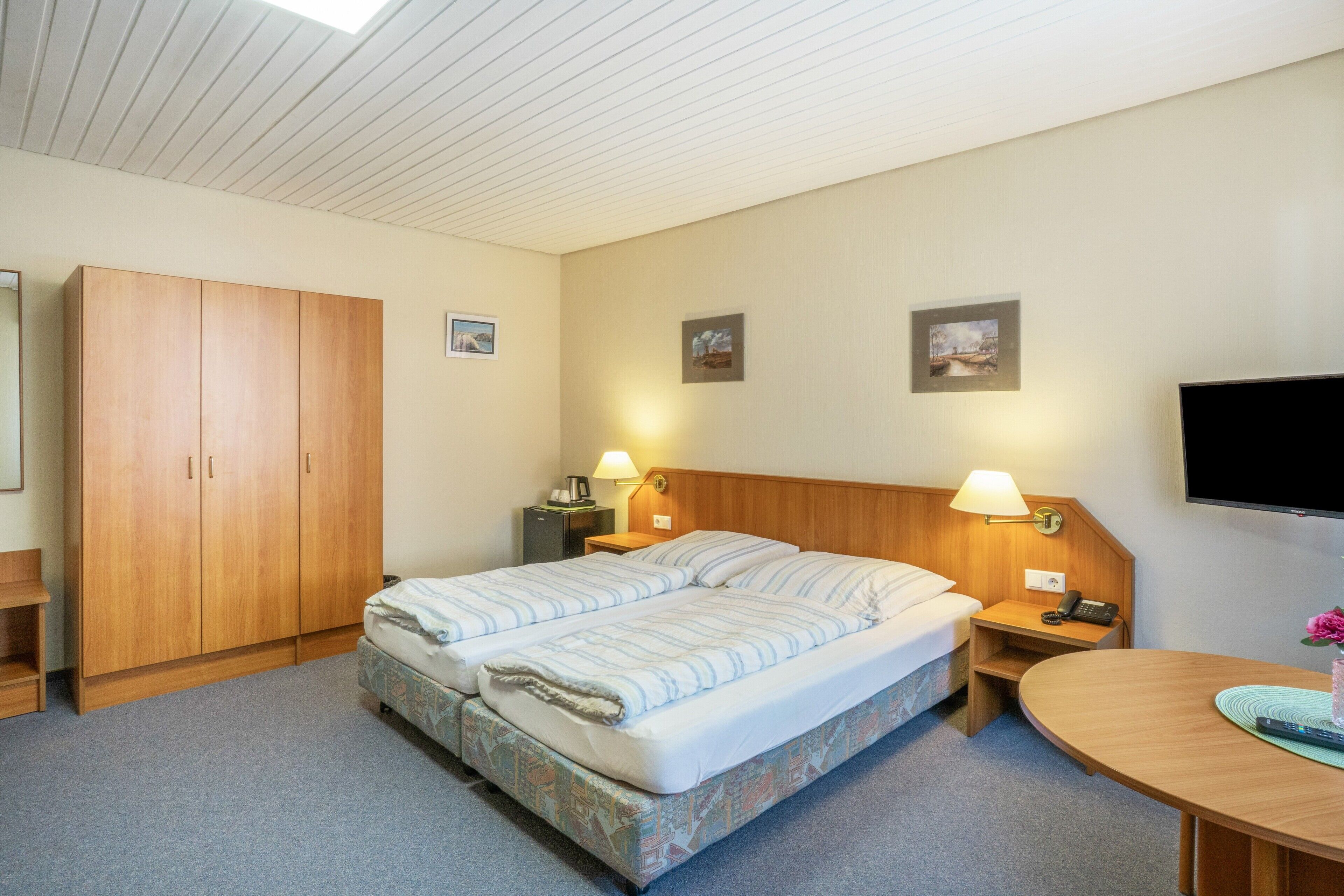 Una habitación de hotel con cama, armario, escritorio con lámpara, televisor y obras de arte en las paredes.