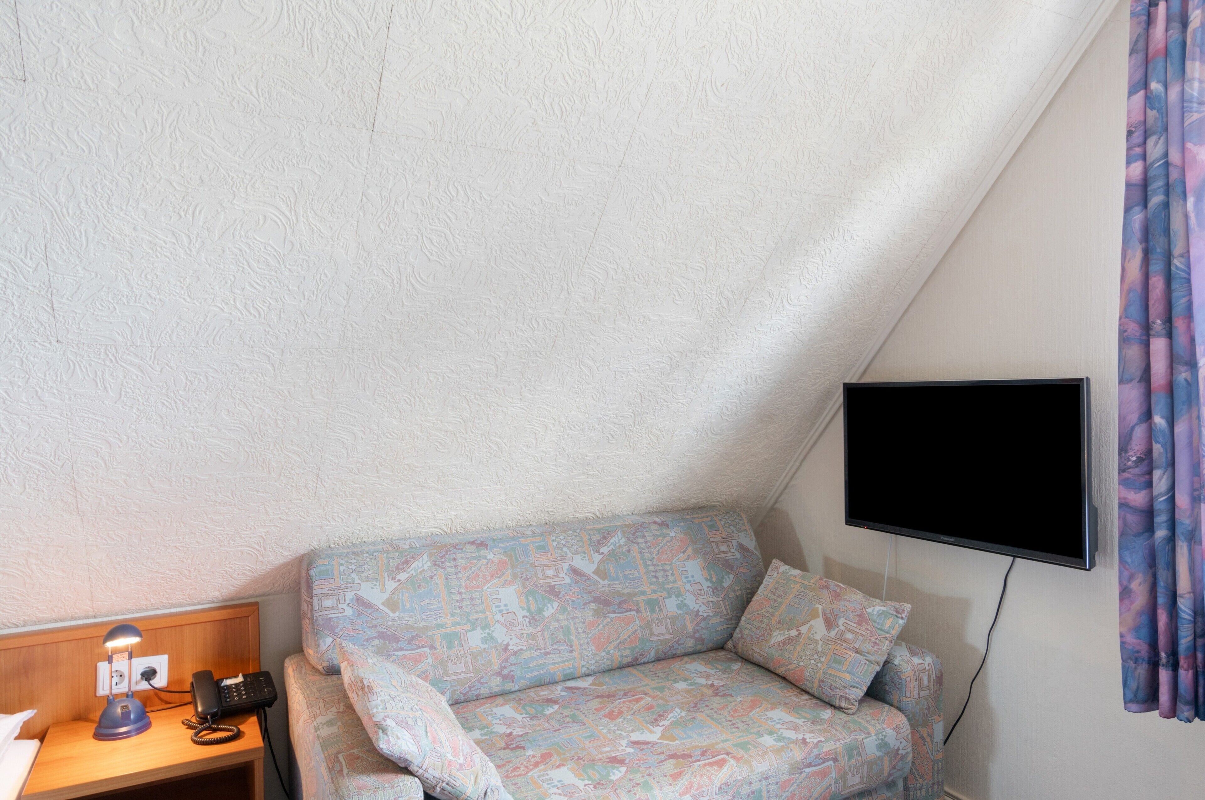 Ein kleiner, dreieckiger Raum mit einer Couch, einem Schreibtisch mit Lampe und einem an der Wand befestigten Flachbildfernseher.