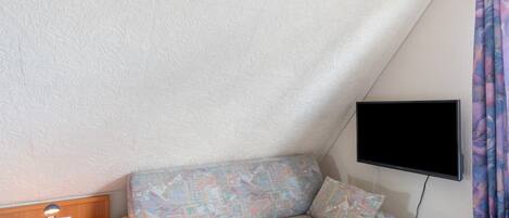 Ein kleiner, dreieckiger Raum mit einer Couch, einem Schreibtisch mit Lampe und einem an der Wand befestigten Flachbildfernseher.