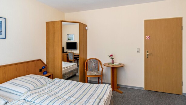A hotel room with a bed, a chair, a small table with a vase, and a door with a no-smoking sign.