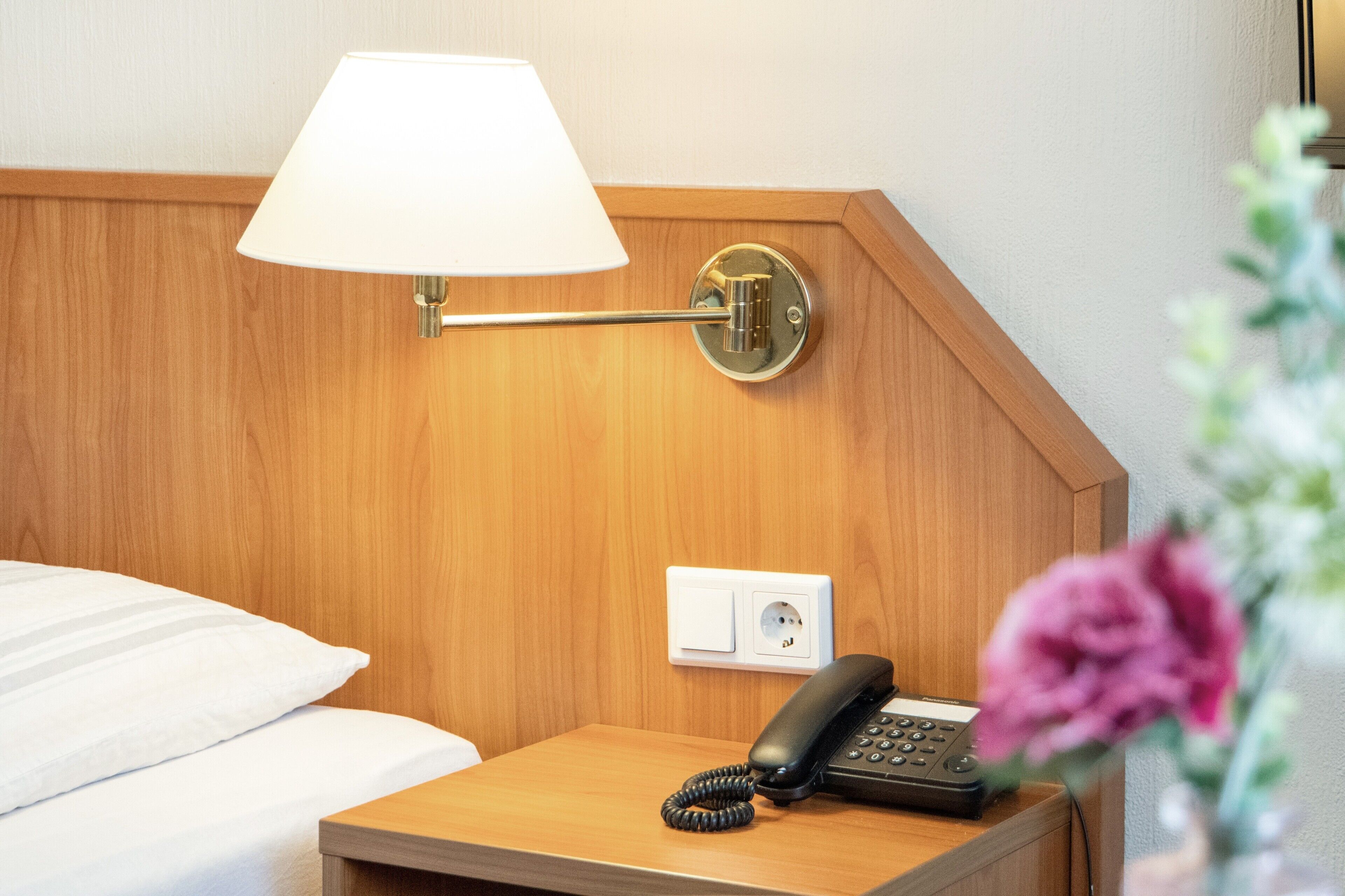 A hotel room with a wooden headboard, a bedside lamp, a telephone, and a power outlet.