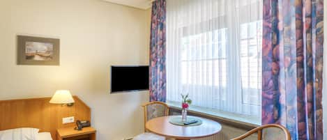 A hotel room with a bed, a desk with a lamp, a television, a window with curtains, and a chair.