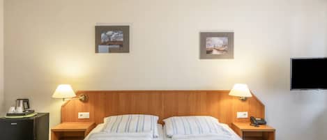 Habitación de hotel con dos camas, mesitas de noche, lámparas, un televisor de pantalla plana y dos cuadros en la pared.