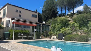 En swimmingpool med stenplattor, en täckt utomhusplats med sittmöbler och en byggnad med ett tak täckt av plattor.
