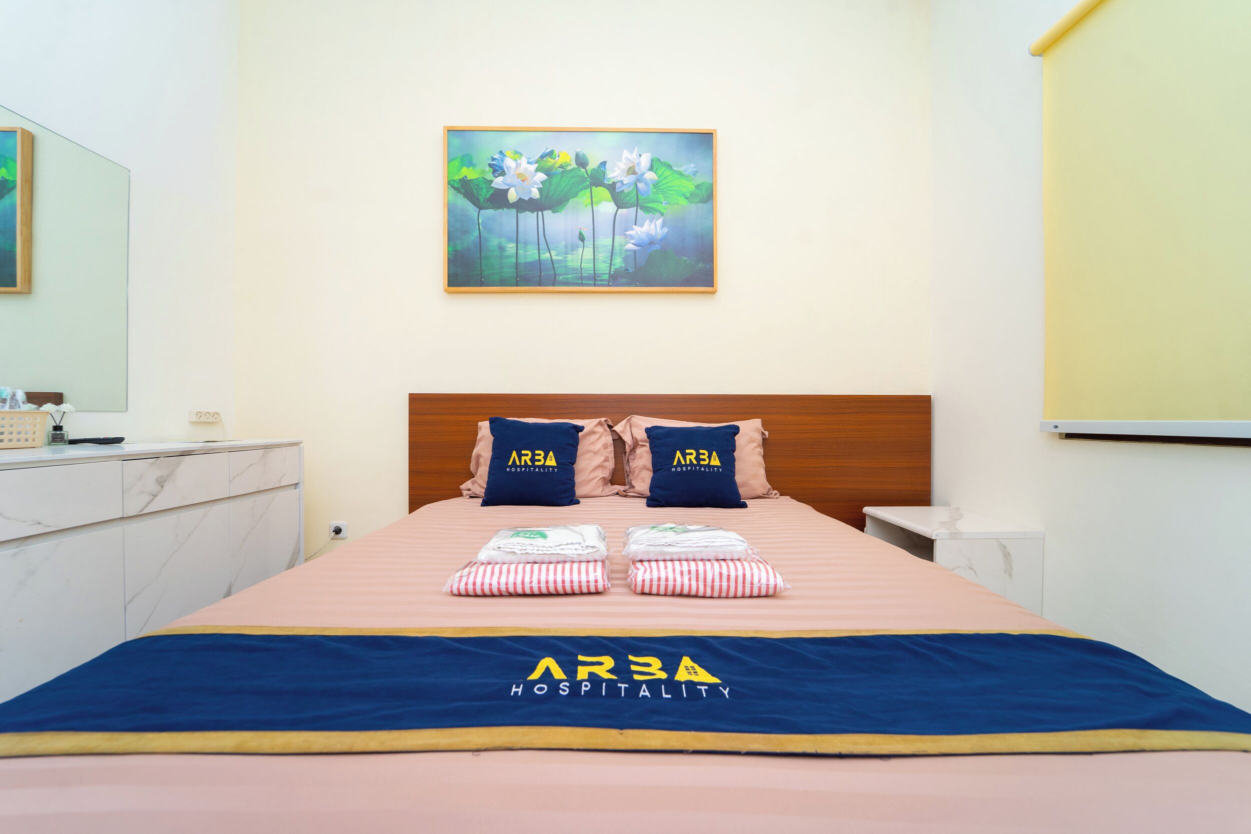 Una habitación de hotel con una cama, almohadas, un cuadro, una mesita de noche y un cabecero con el nombre del hotel.