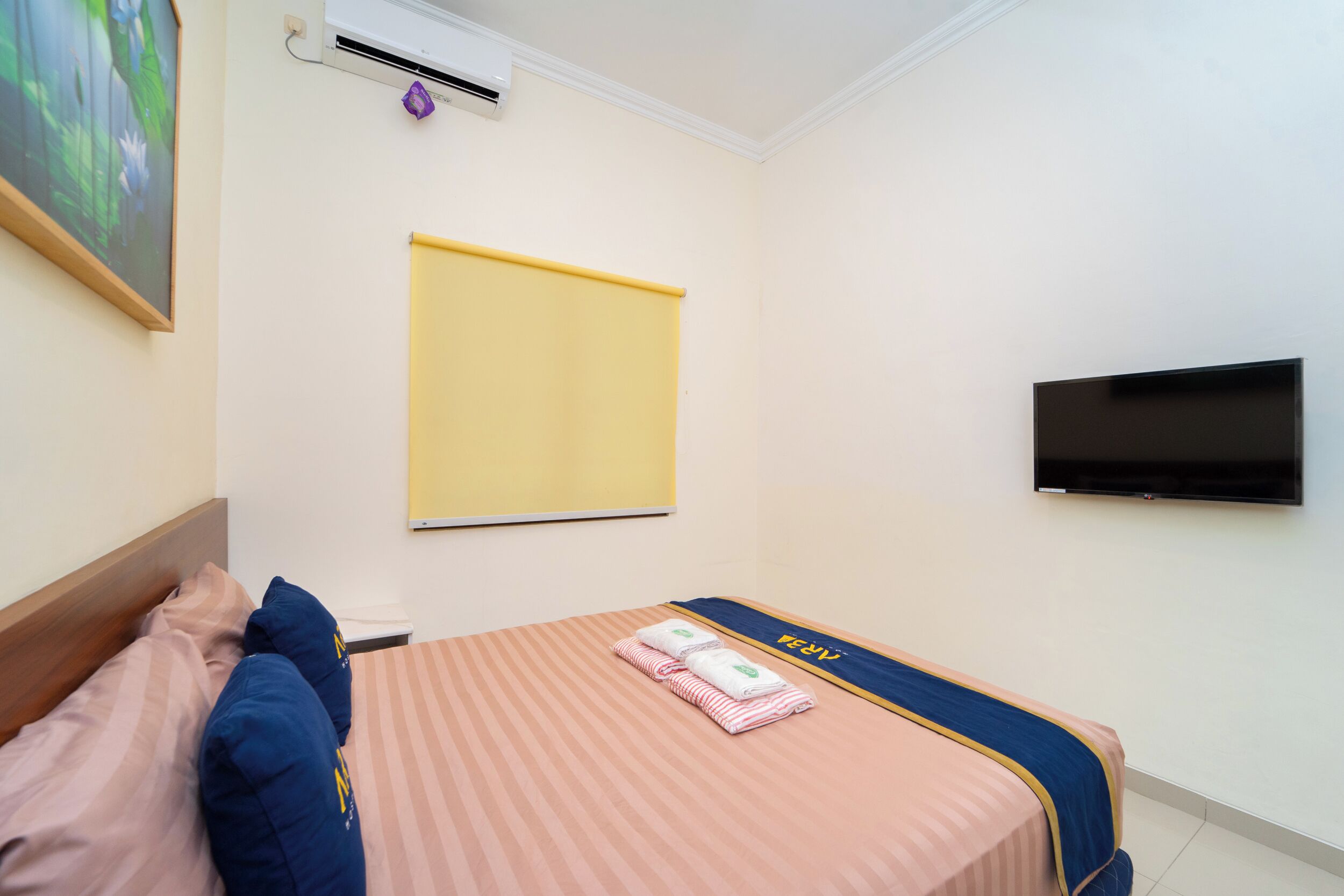 A hotel room with a bed, a flat-screen TV, an air conditioning unit, and a painting on the wall.