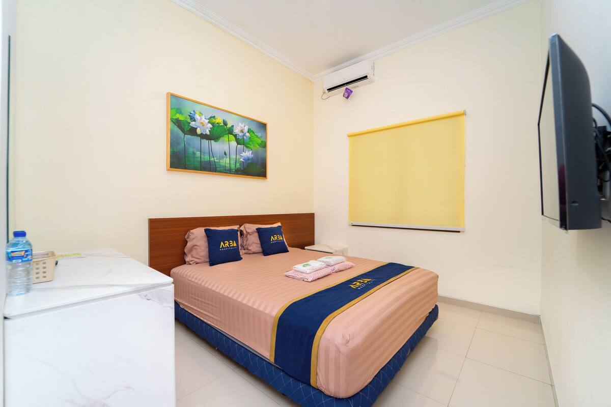 A hotel room with a bed, a desk with a water bottle, a painting, an air conditioner, and a TV.