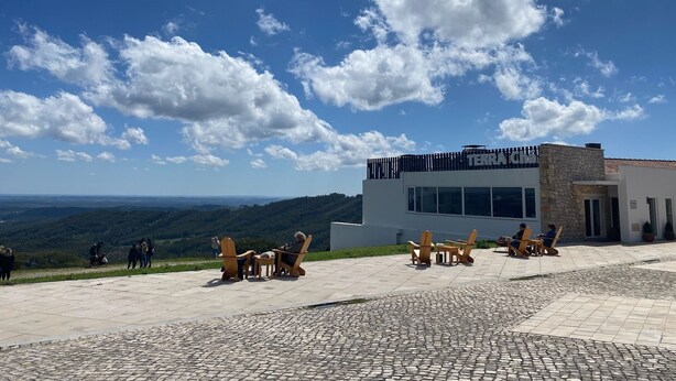 A modern building with a stone facade and a flat roof, surrounded by a paved patio area with wooden chairs and tables. - 2 bedroom house in the heart of the Serra d'Aire e Candeeiros natural park (Rio Maior)