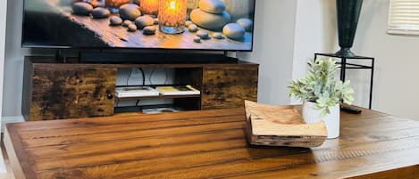 A wooden dining table with a television displaying a candlelit scene, a potted plant, and a decorative wooden tray with letters.