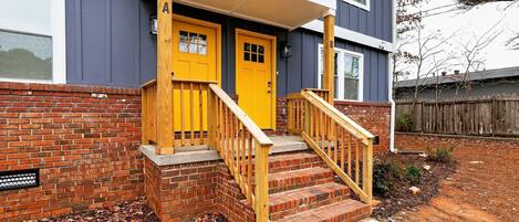 A two-story house with a yellow front door, a brick exterior, and a wooden staircase leading to the entrance.
