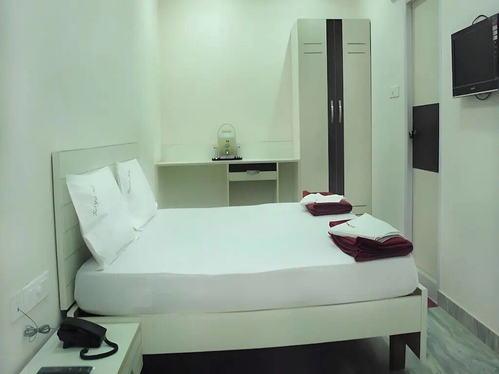 Habitación de hotel con cama, teléfono en la mesita de noche, armario, escritorio con lámpara y televisor en la pared.