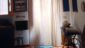Una habitación con techo de madera, una ventana con cortina, un estante en la pared con utensilios de cocina, una mesa pequeña con una silla y una máquina de coser.